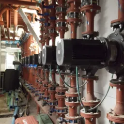 Szereg czerwonych rur instalacyjnych z zaworami i czarnymi silnikami pomp, widoczne skrzynki narzędziowe na podłodze w tle