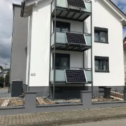 Trzypiętrowy budynek z balkonami z metalową konstrukcją i szklanymi panelami, na których zamontowano panele słoneczne; widoczny numer domu '40' i ogrodzenie z metalową siatką.