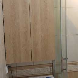 Paweł Kiliszek - Szafka wisząca z drewnopodobnymi frontami, zamontowana nad białą pralką Samsung w łazience z beżowymi płytkami i kabiną prysznicową.
