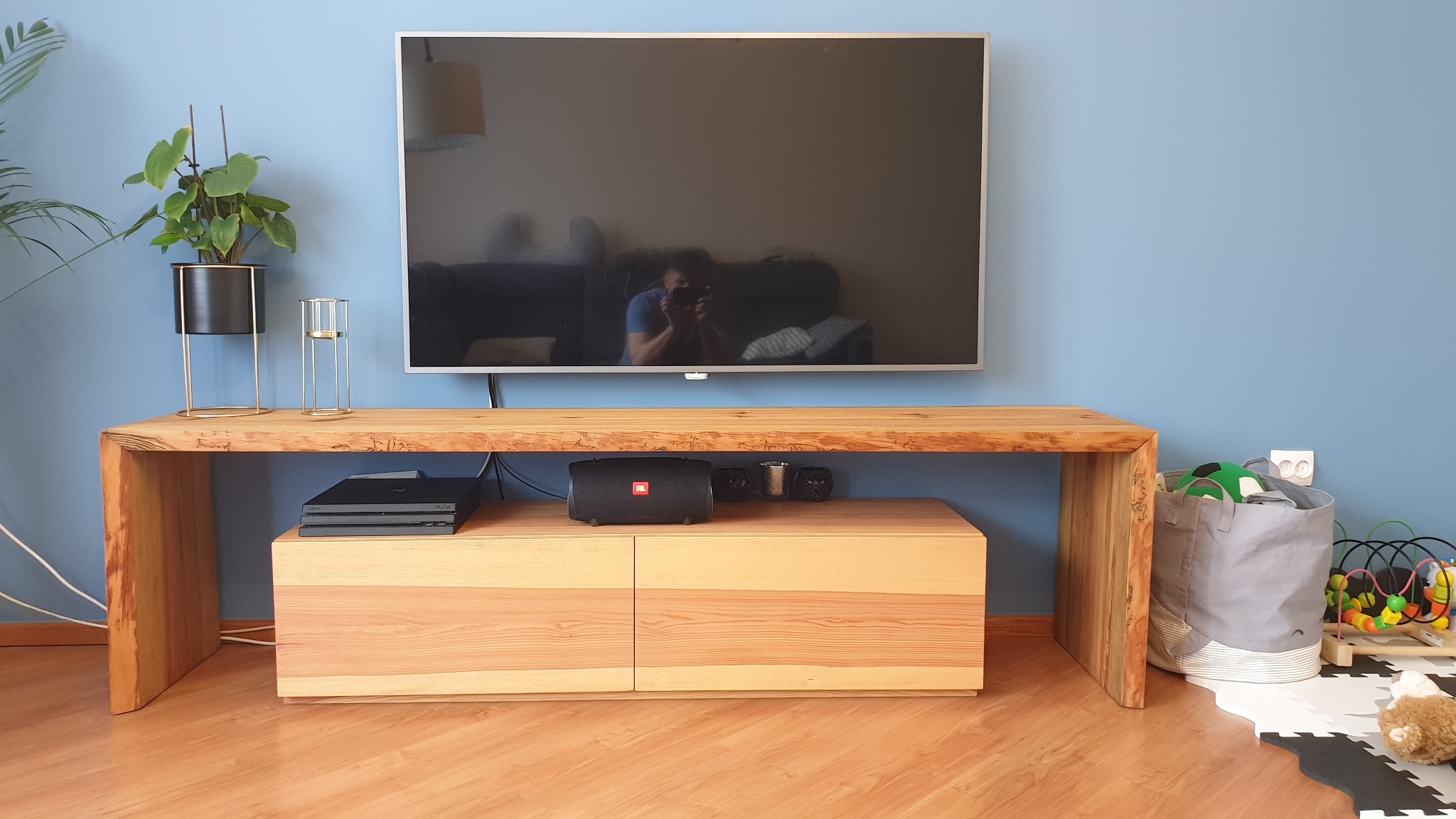 Drewniana szafka RTV z litego drewna z dwoma szufladami, na niej telewizor odbijający fotografa, konsola do gier, głośnik JBL i dekoracje, obok kosz na zabawki, ściana pomalowana na niebiesko.