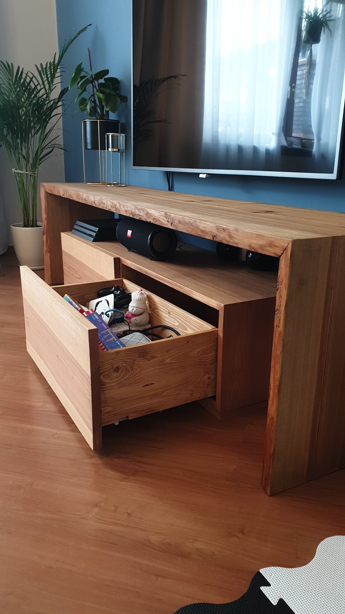Drewniana szafka RTV z wysuwaną szufladą, w której widać różne przedmioty, w tle telewizor i rośliny doniczkowe.