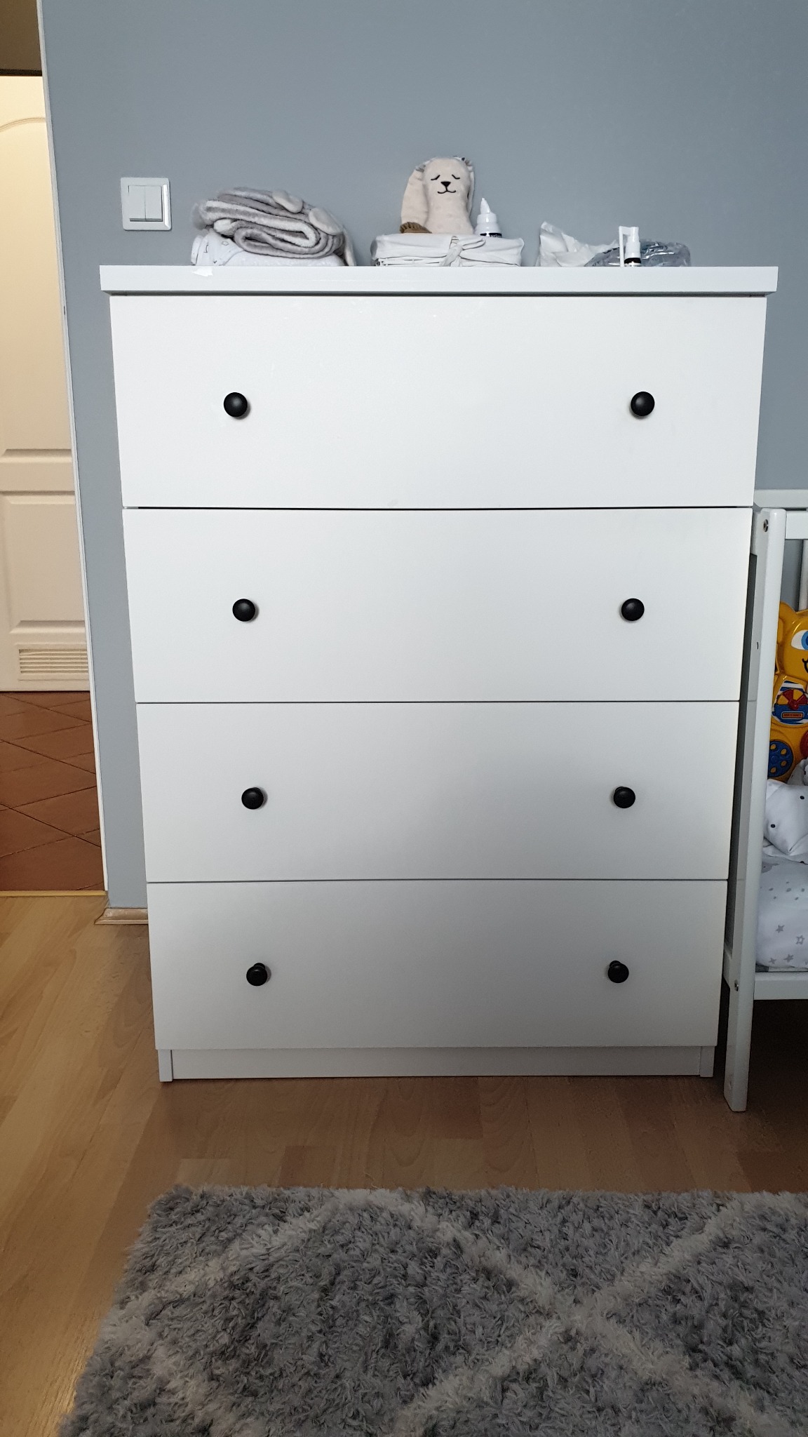 Biała komoda z czterema szufladami i czarnymi okrągłymi uchwytami, na blacie ułożone dziecięce akcesoria i pluszowy miś, obok widoczne łóżeczko dziecięce, na podłodze szary dywan.