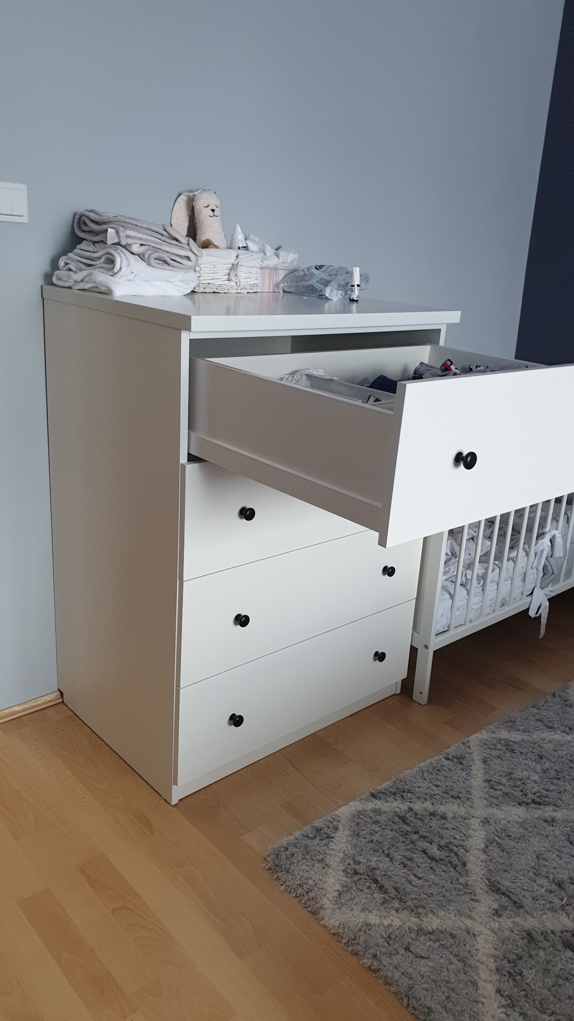 Biała komoda z czterema szufladami, jedna otwarta, czarne okrągłe uchwyty, na blacie ułożone ubranka i pluszowy królik, obok białe łóżeczko dziecięce, na podłodze szary dywan.