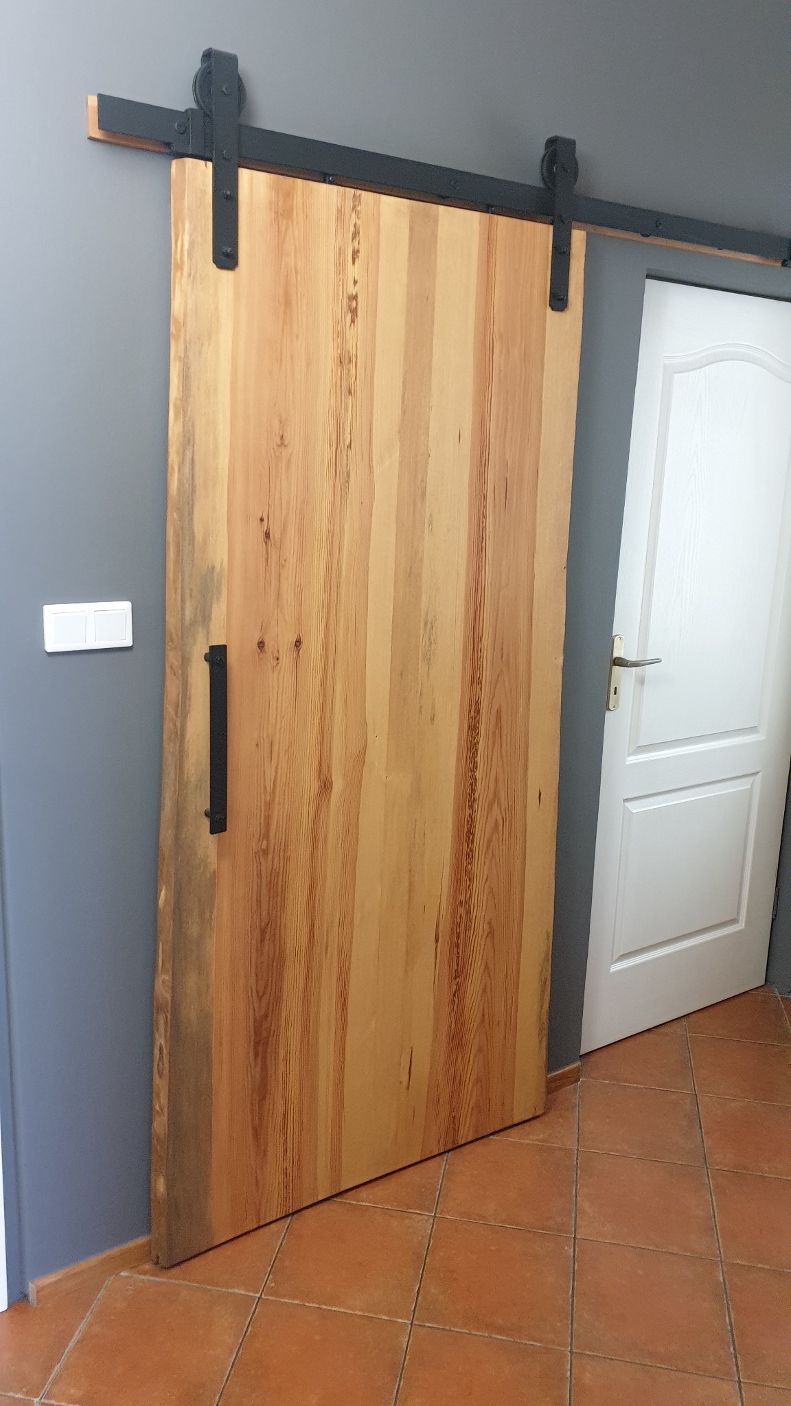 Drewniane drzwi przesuwne typu stodoła z czarnym okuciem i uchwytem, widoczne na tle szarej ściany i białych drzwi.