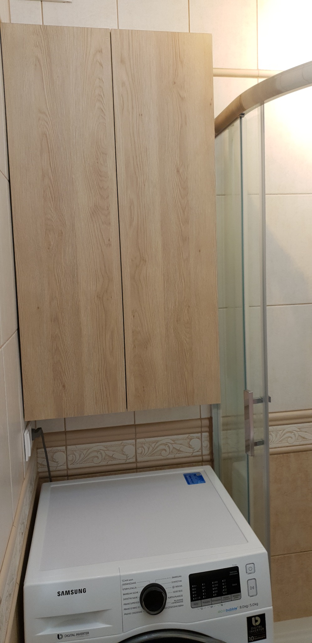 Szafka wisząca z drewnopodobnymi frontami, zamontowana nad białą pralką Samsung w łazience z beżowymi płytkami i kabiną prysznicową.