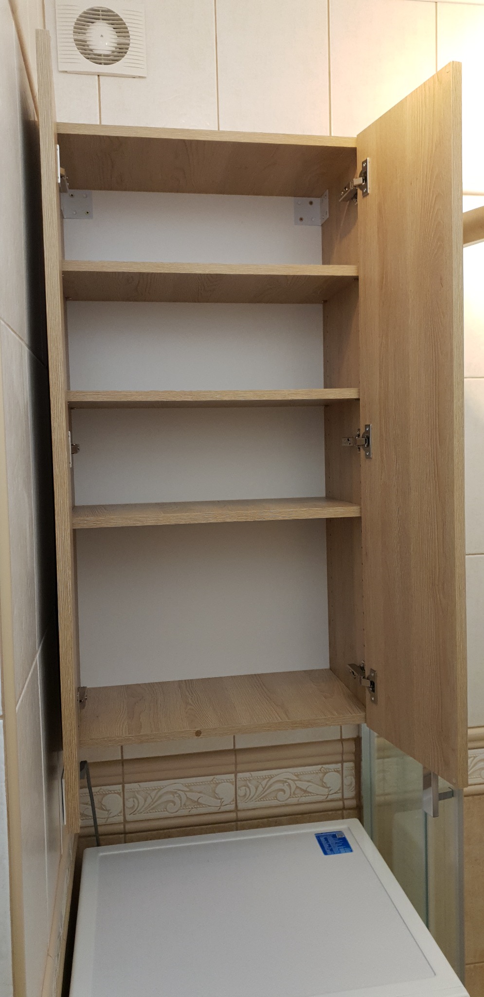 Otwarta szafka łazienkowa z półkami o strukturze jasnego drewna, zamontowana nad białą pralką w łazience wyłożonej beżowymi płytkami z dekoracyjnym wzorem.