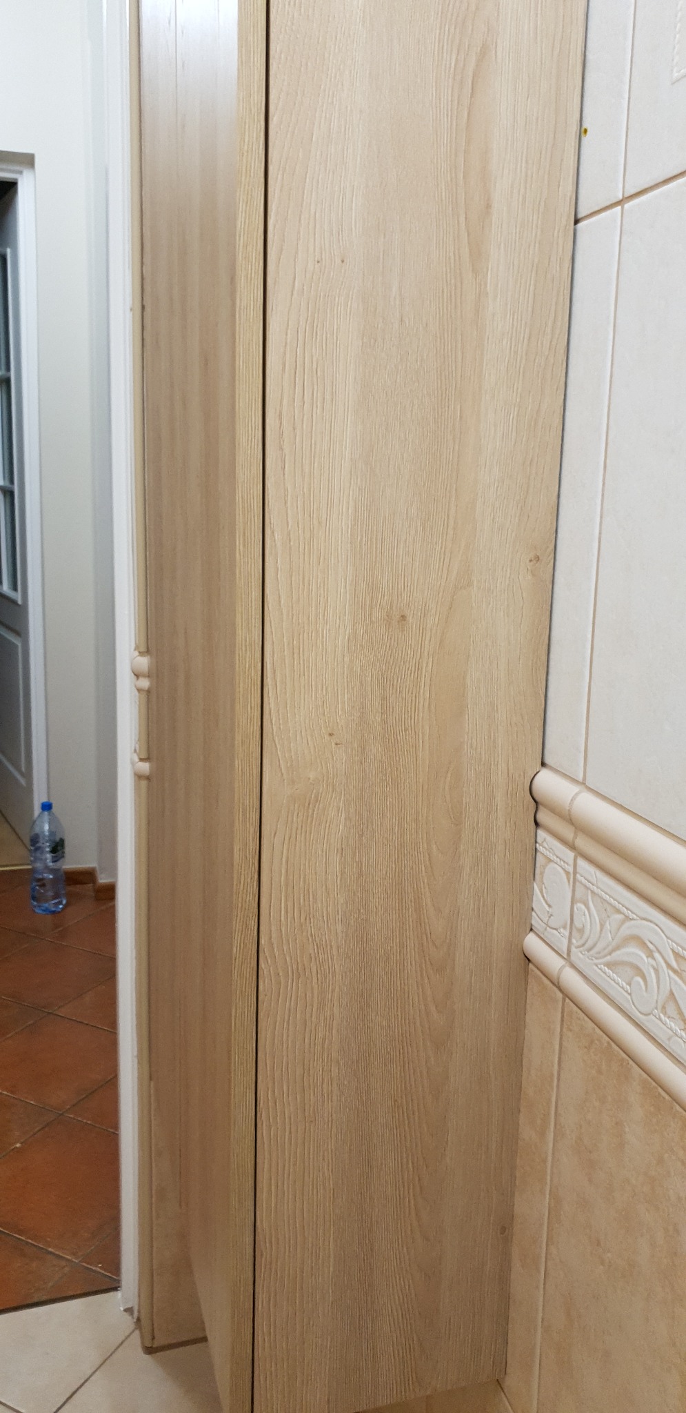 Bliski kadr na zamknięte, dwudrzwiowe szafy z jasnego drewna z widoczną fakturą słojów, umieszczone w rogu pomieszczenia z beżowymi płytkami i dekoracyjnym pasem ceramicznym, butelka wody w tle.