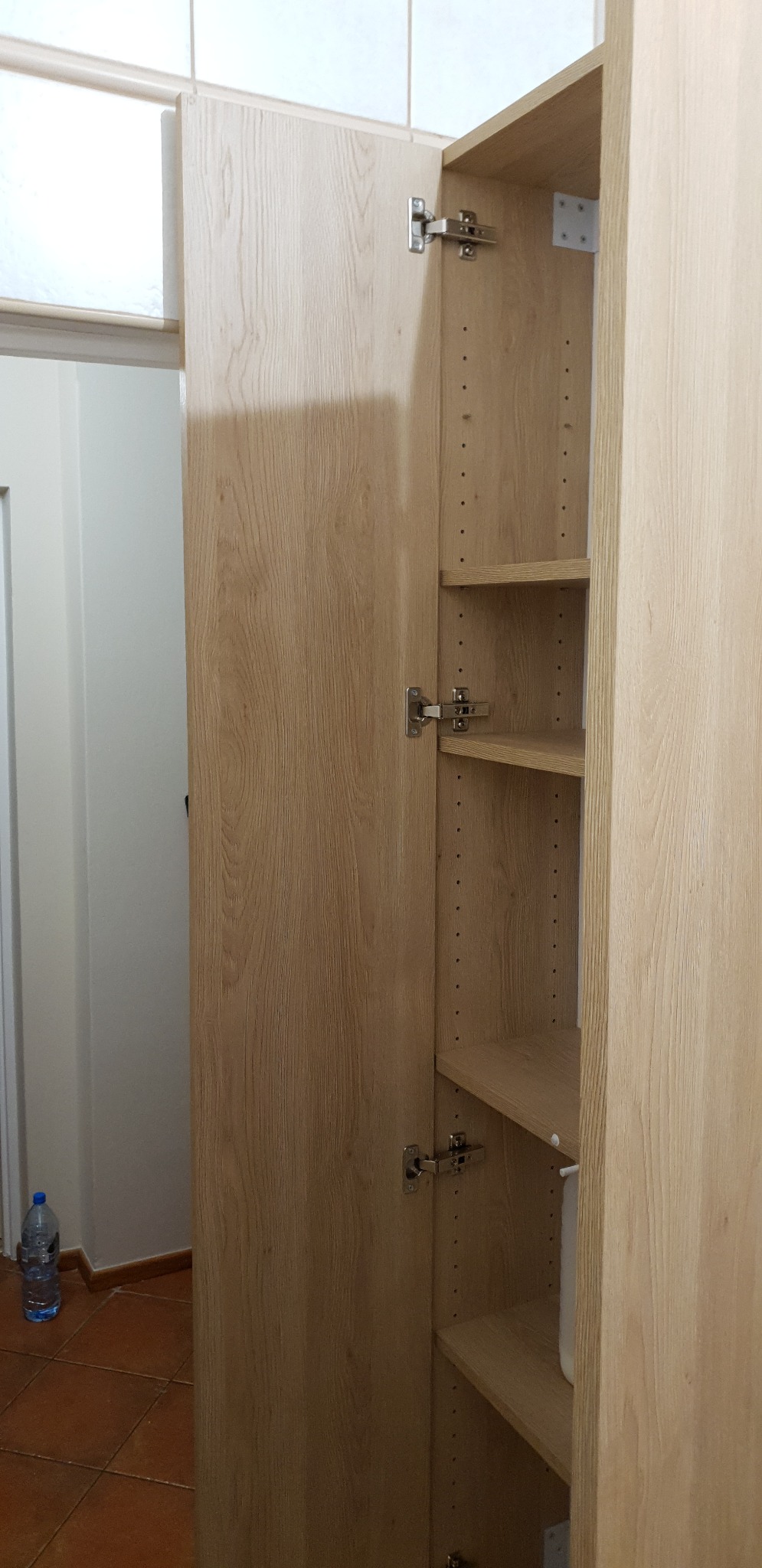 Otwarta szafa z jasnego drewna z regulowanymi półkami, widoczne zawiasy i nawiercone otwory na różnych wysokościach, butelka wody na podłodze w tle.