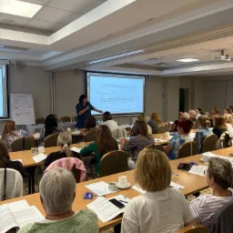Sala konferencyjna podczas szkolenia: prelegentka omawia prezentację na rzutniku, uczestnicy siedzą przy stołach z notatkami i napojami. Widoczny flipchart.