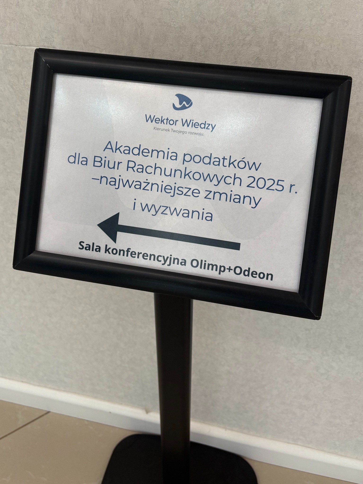 Tablica informacyjna z napisem 'Akademia podatków dla Biur Rachunkowych 2025' i strzałką wskazującą kierunek do Sali konferencyjnej Olimp+Odeon.