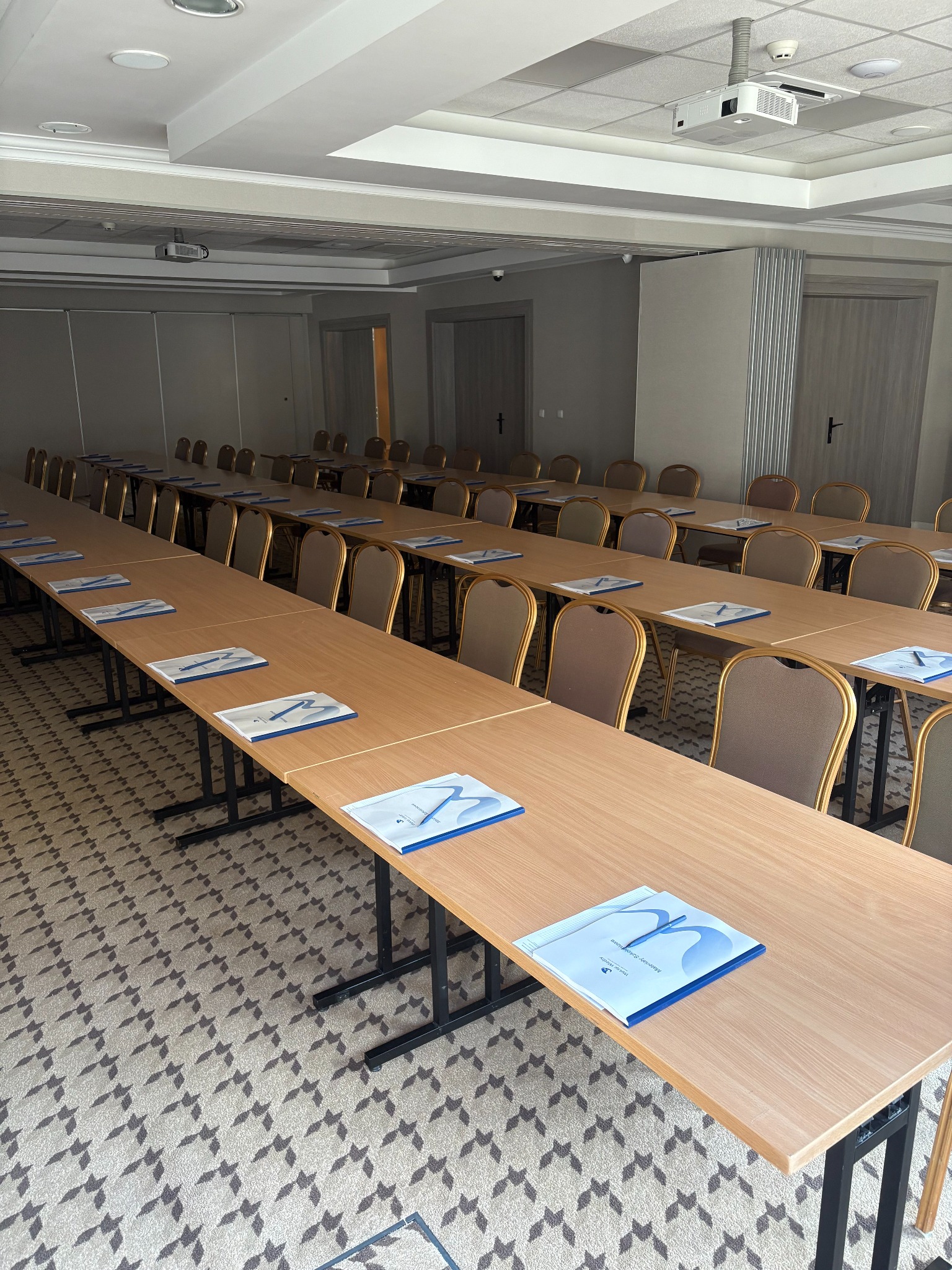 Sala konferencyjna z długimi stołami, krzesłami i materiałami szkoleniowymi. Projektor sufitowy. Wzór na wykładzinie. Przygotowane miejsce na szkolenie.