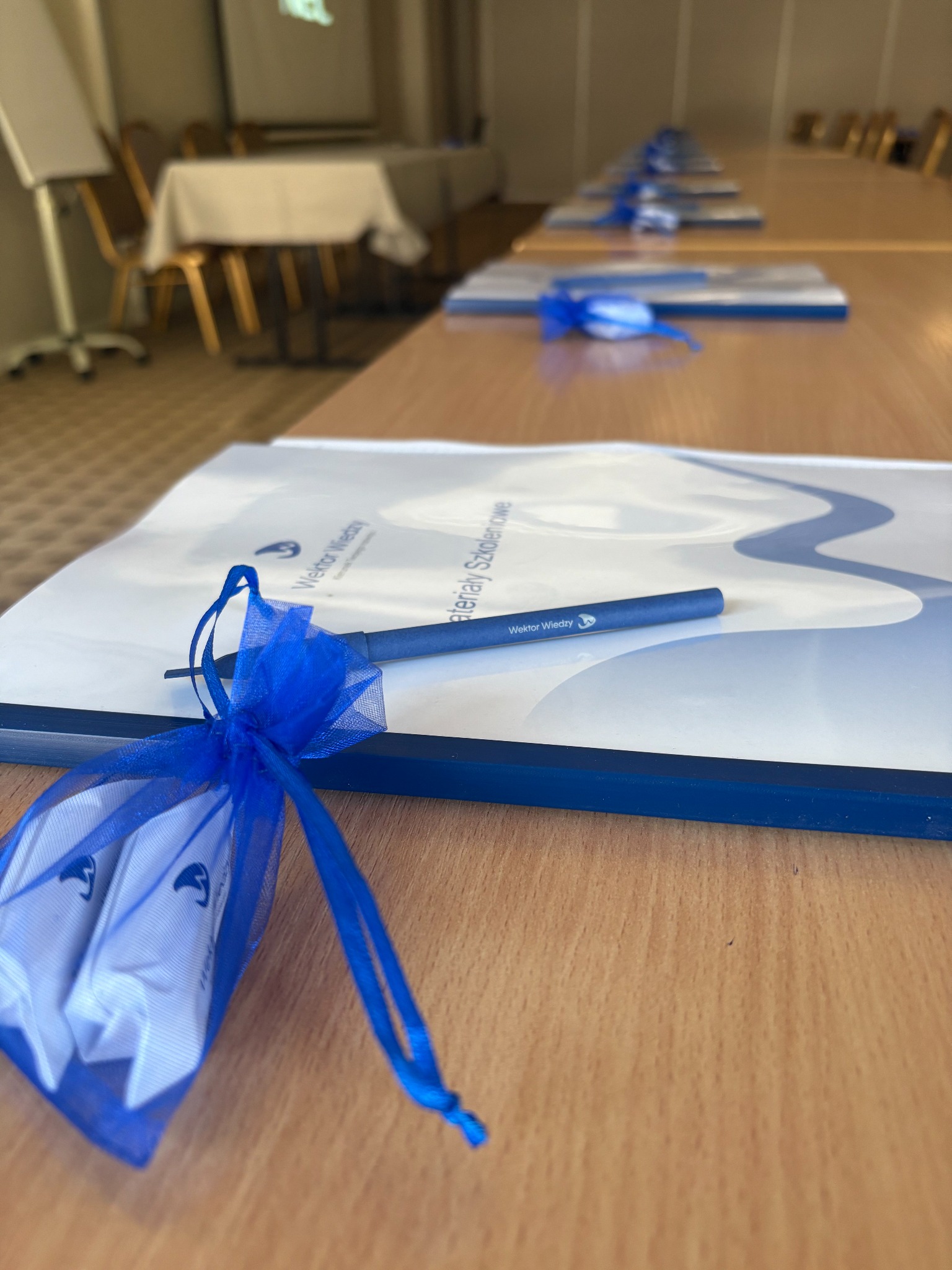 Sala konferencyjna: materiały szkoleniowe, długopis i upominki w niebieskich woreczkach na drewnianym stole. W tle krzesła i rzutnik.