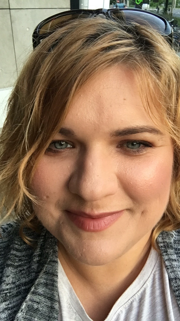 Portret kobiety z blond włosami i niebieskimi oczami, w okularach przeciwsłonecznych na głowie, uśmiechająca się delikatnie.