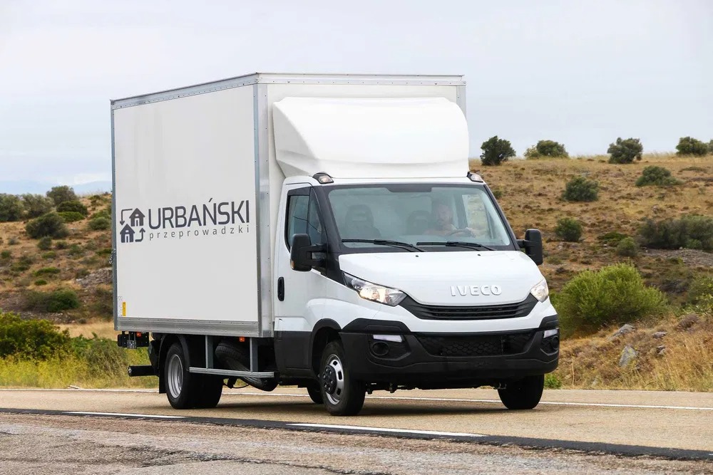 Biały samochód dostawczy Iveco z nadrukowanym logo firmy przeprowadzkowej 'Urbański' jadący po asfaltowej drodze, w tle pagórkowaty krajobraz.