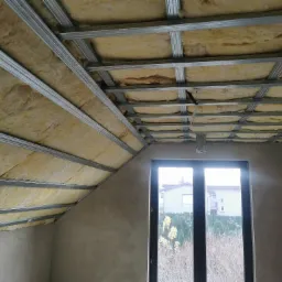 Poddasze z widoczną izolacją z wełny mineralnej umieszczoną między metalowymi profilami konstrukcyjnymi sufitu podwieszanego, okno w tle.