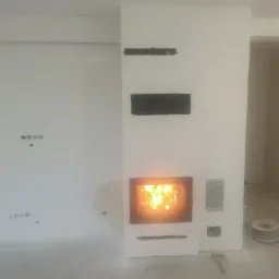 Wnętrze w trakcie wykańczania z wbudowanym kominkiem z palącym się ogniem, widoczne niedoskonałości na ścianach i instalacja elektryczna.