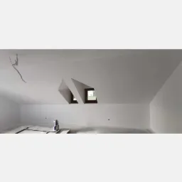 Poddasze po wykonaniu prac wykończeniowych, widoczne białe ściany, okna dachowe i przygotowane podłoże pod dalsze prace, butelka z rozpylaczem i wałek malarski na podłodze.