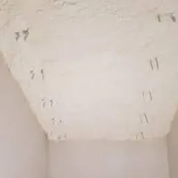 Nierównomiernie nałożona, jasna warstwa izolacyjna z widocznymi fragmentami mocowania na skośnej powierzchni poddasza w trakcie prac wykończeniowych.