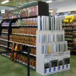 Wnętrze sklepu budowlanego: regały z farbami, lakierami, profilami aluminiowymi i innymi materiałami wykończeniowymi. Widoczny szyld 'BOTAMENT'.