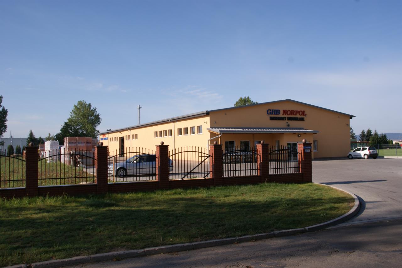 Żółty budynek składu budowlanego GHB NORPOL z zaparkowanymi autami, ogrodzony niskim płotem z cegły i metalu, w tle składowane materiały budowlane.