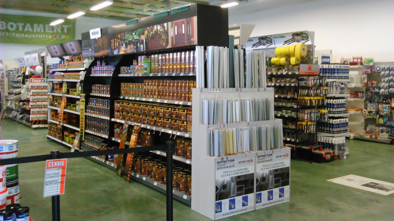 Wnętrze sklepu budowlanego: regały z farbami, lakierami, profilami aluminiowymi i innymi materiałami wykończeniowymi. Widoczny szyld 'BOTAMENT'.