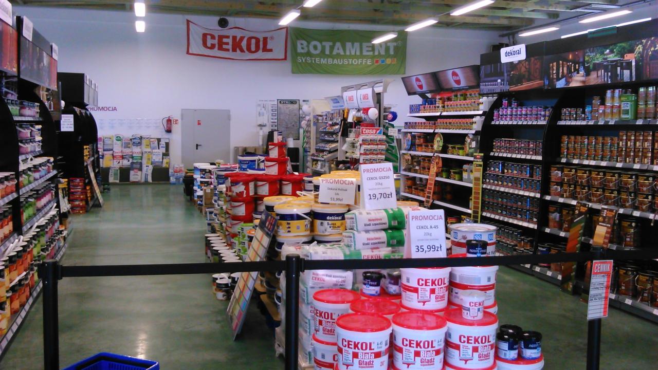 Wnętrze sklepu budowlanego: regały z farbami, lakierami i chemią budowlaną. Widoczne promocje na produkty marek Cekol i Dekoral. Uwagę przyciągają palety z wiadrami białej gładzi.