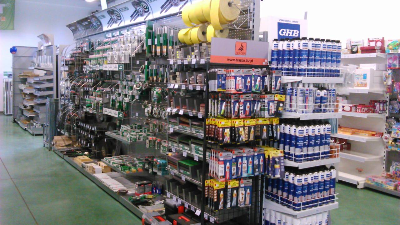 Wnętrze sklepu budowlanego: regały z narzędziami, klejami, uszczelniaczami i innymi materiałami. Widoczne opakowania marki Dragon i GHB.