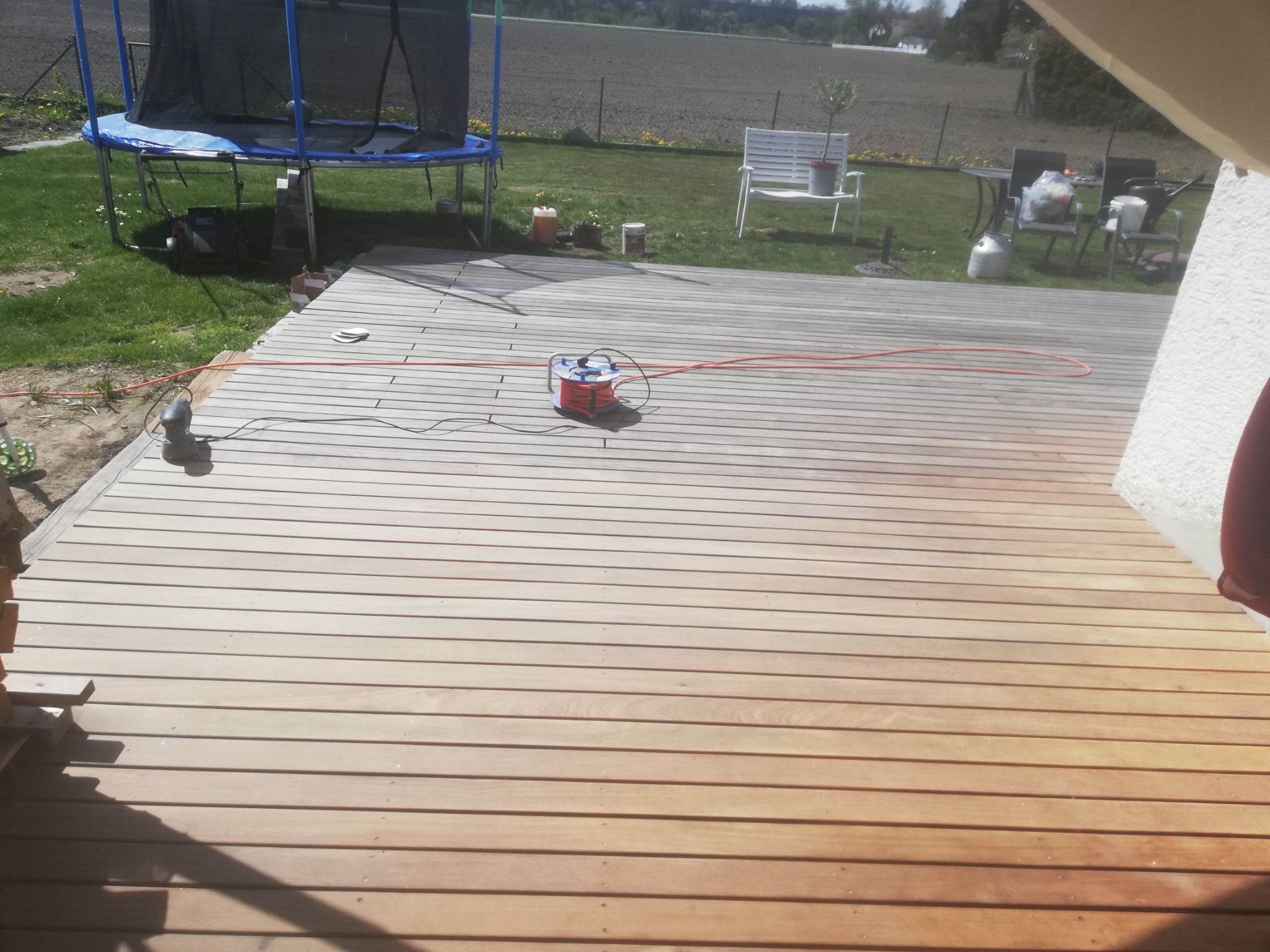 Świeżo pomalowany drewniany taras z widocznym zwiniętym przedłużaczem i pistoletem natryskowym, w tle trampolina i meble ogrodowe.