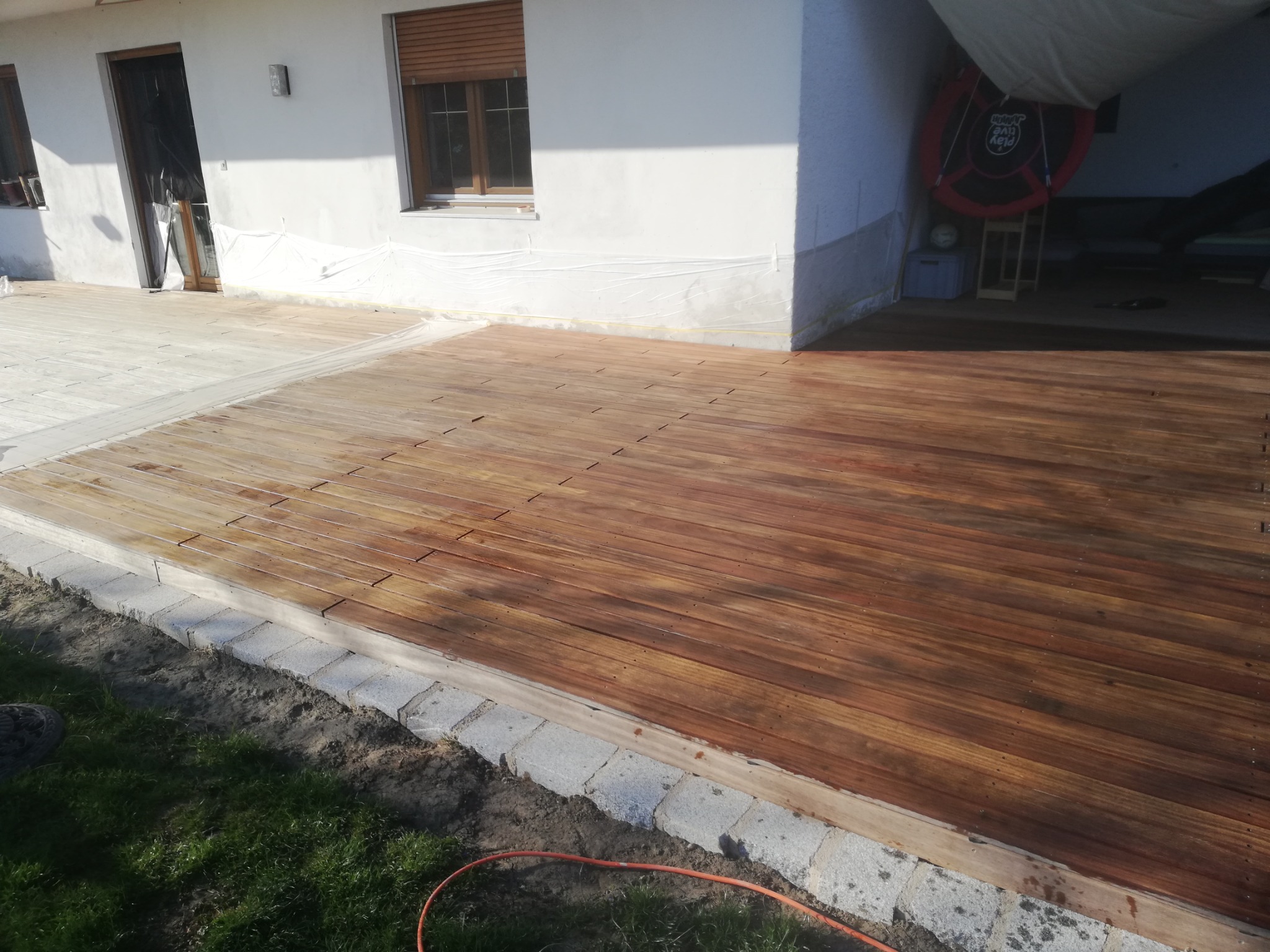 Nowa, drewniana deska tarasowa z widocznymi śladami świeżego lakieru, obłożona szarą kostką brukową, przy białym budynku z oknami zabezpieczonymi folią malarską.