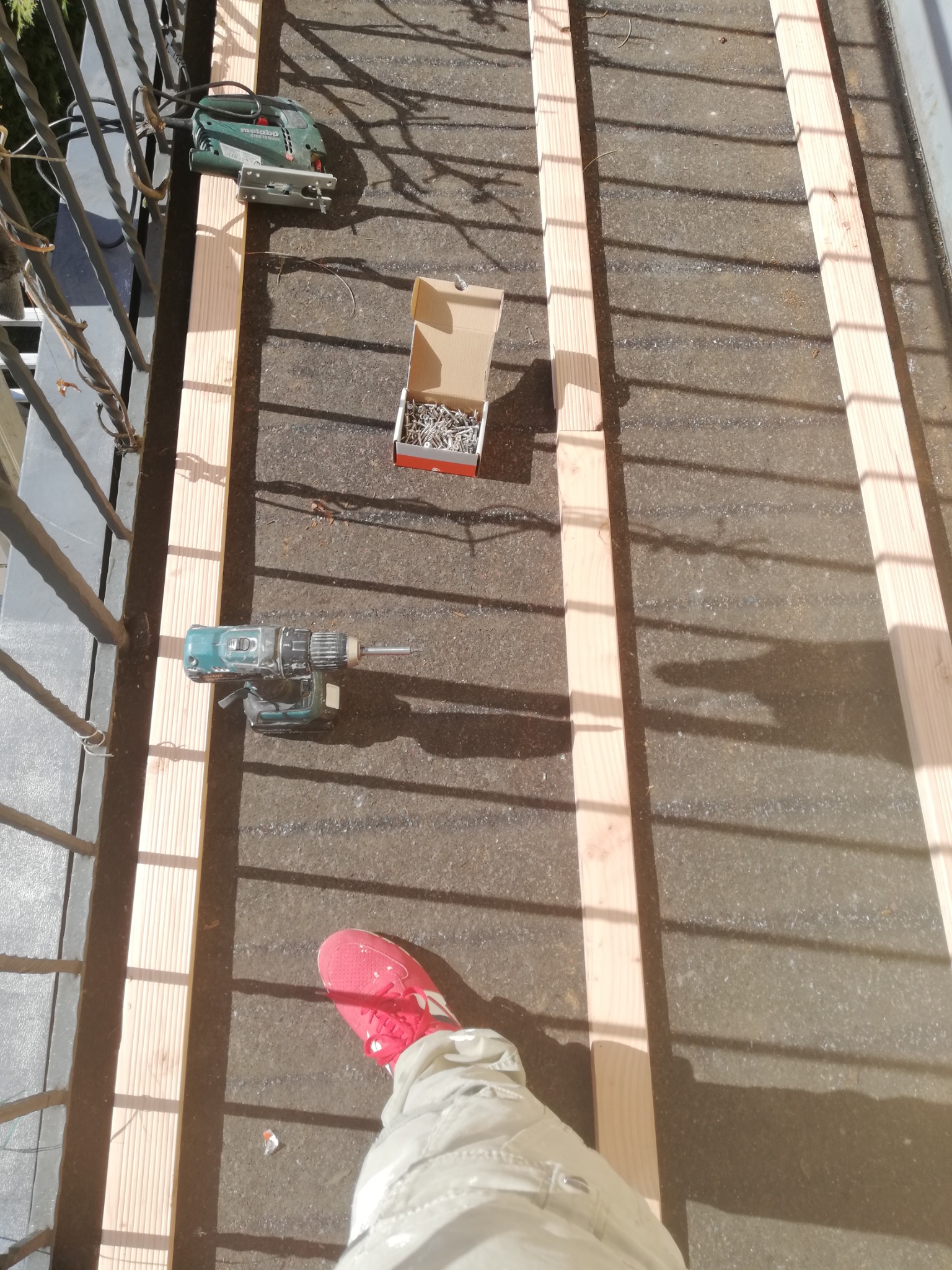 Montaż drewnianej konstrukcji na balkonie: deski, wkrętarka, wyrzynarka i pudełko gwoździ w promieniach słońca, widok z góry na fragment nogi w różowym bucie.