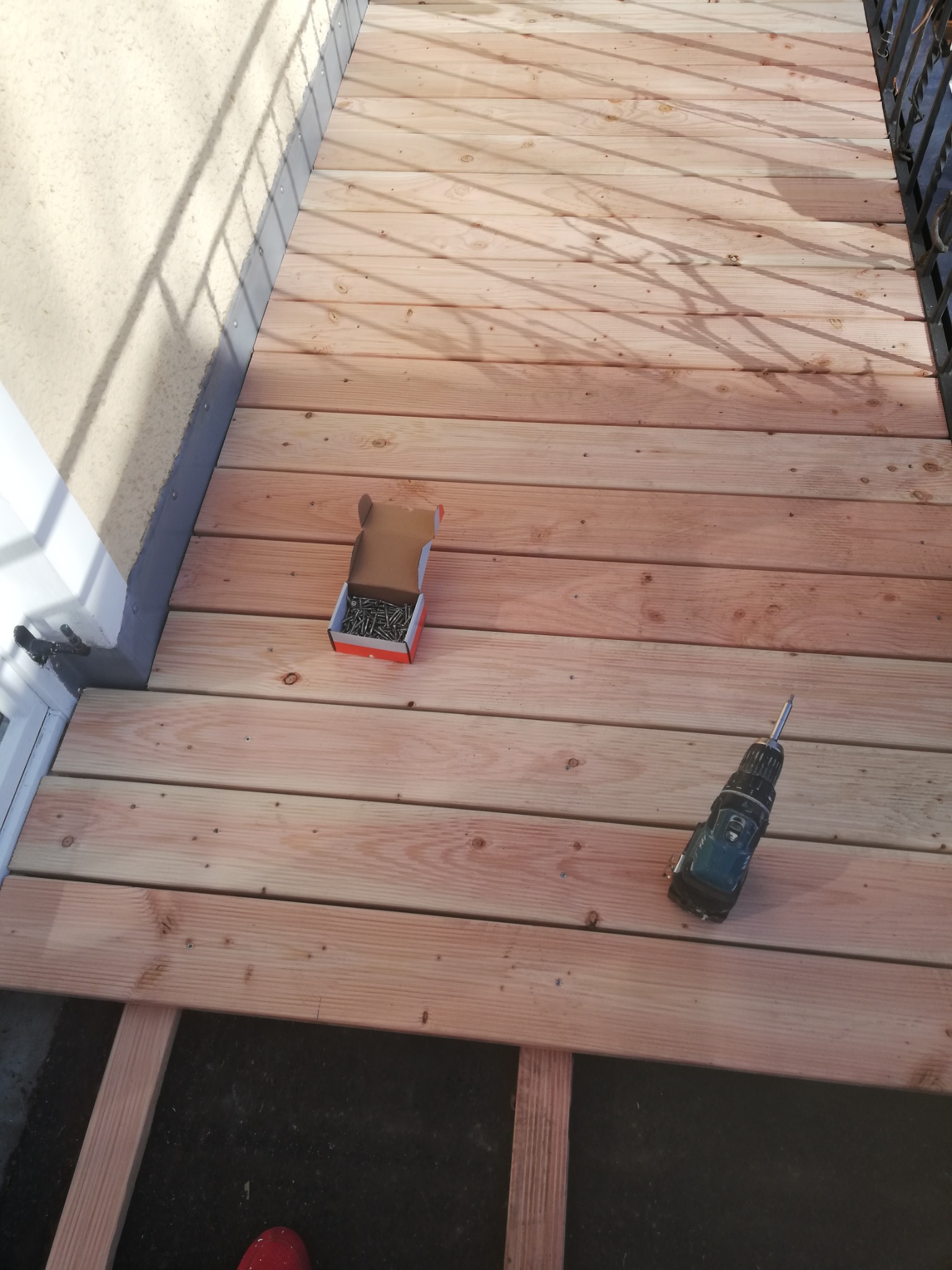 Montaż drewnianej podłogi na balkonie: widoczne deski, wkrętarka i pudełko z wkrętami, częściowo ukończona konstrukcja.