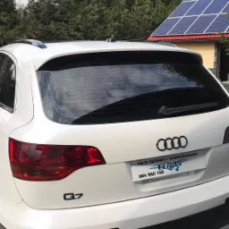 Białe Audi Q7 z przyciemnionymi tylnymi szybami, widoczne z tyłu na tle domu z panelami słonecznymi i drzewami.