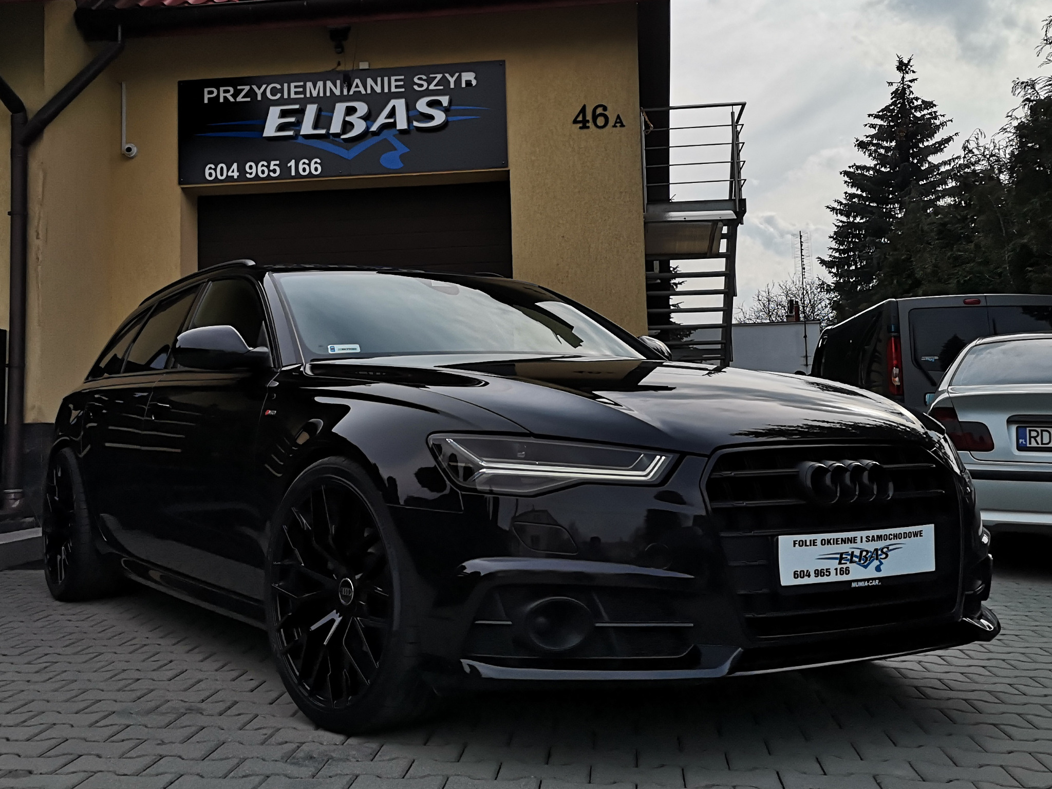 Czarny samochód Audi kombi zaparkowany przed budynkiem z szyldem 'Przyciemnianie szyb ELBAS' i numerem 46A, prezentujący folię okienną i samochodową z numerem telefonu.