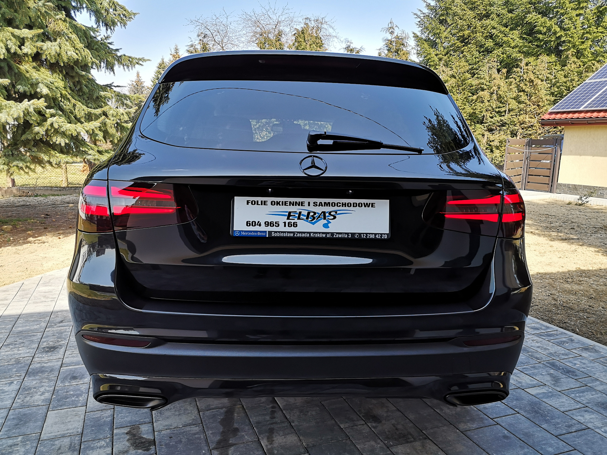 Czarne auto marki Mercedes z przyciemnionymi szybami, widok z tyłu, na tle zieleni w Dębicy. Na tablicy rejestracyjnej naklejka z reklamą firmy.