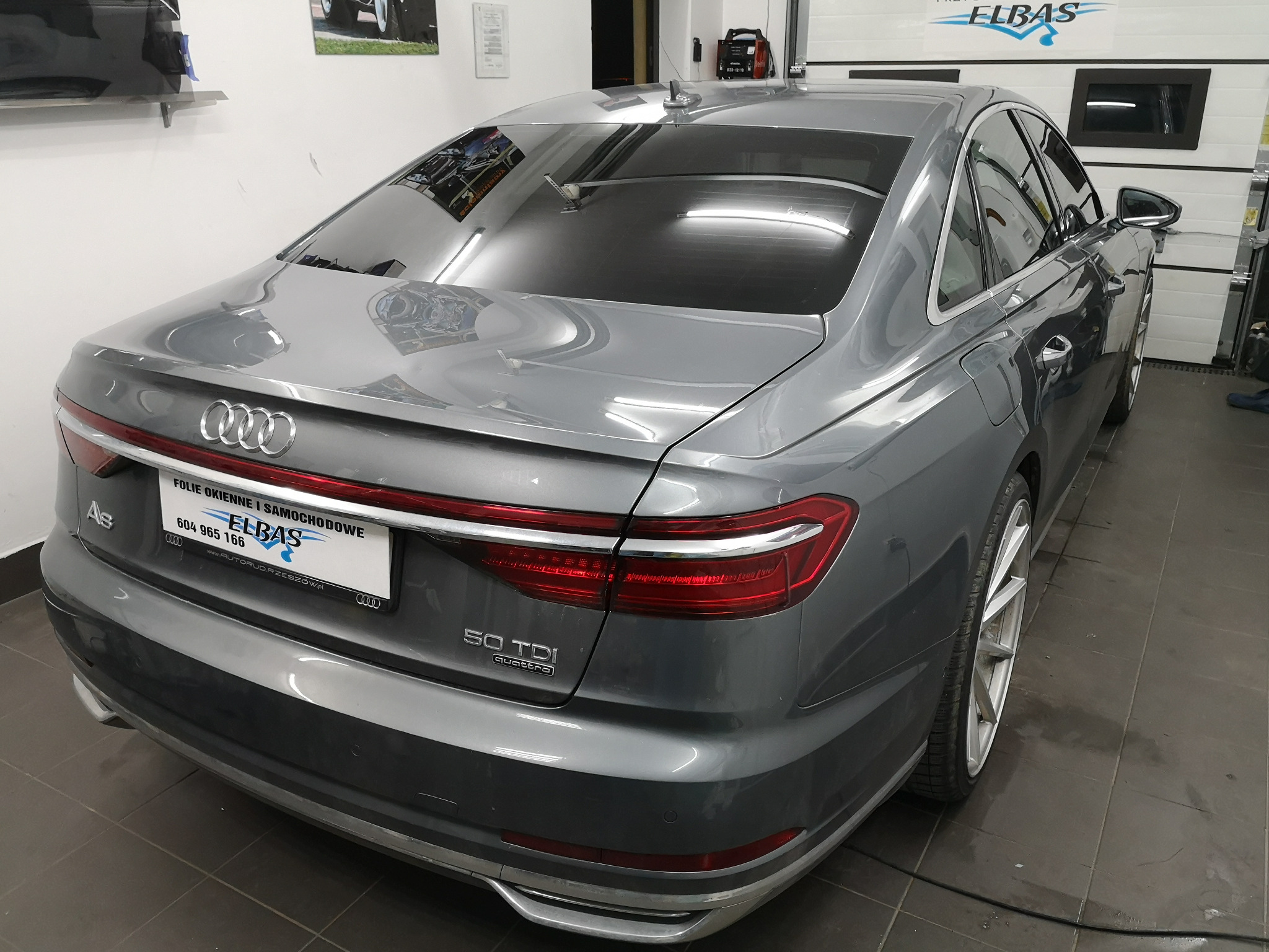 Szary samochód Audi A8 po zabiegu przyciemniania szyb, widoczne ciemne szyby tylne i boczne, tablica rejestracyjna z nazwą firmy 'ELBAS' zajmującej się foliami okiennymi i samochodowymi, widok...