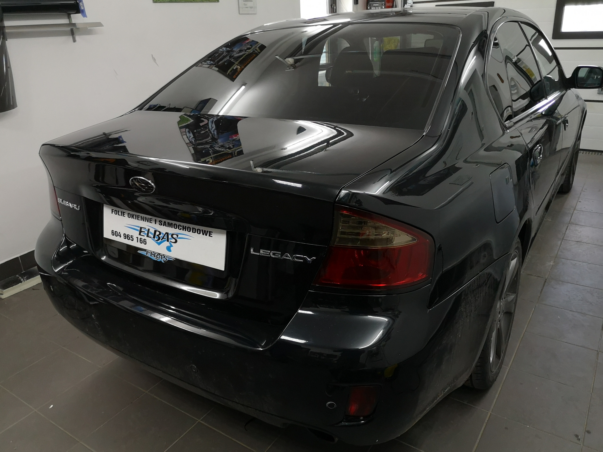 Czarne Subaru Legacy po przyciemnieniu szyb, widoczny tylny fragment pojazdu z mocno przyciemnioną szybą i tablicą rejestracyjną z nazwą firmy.