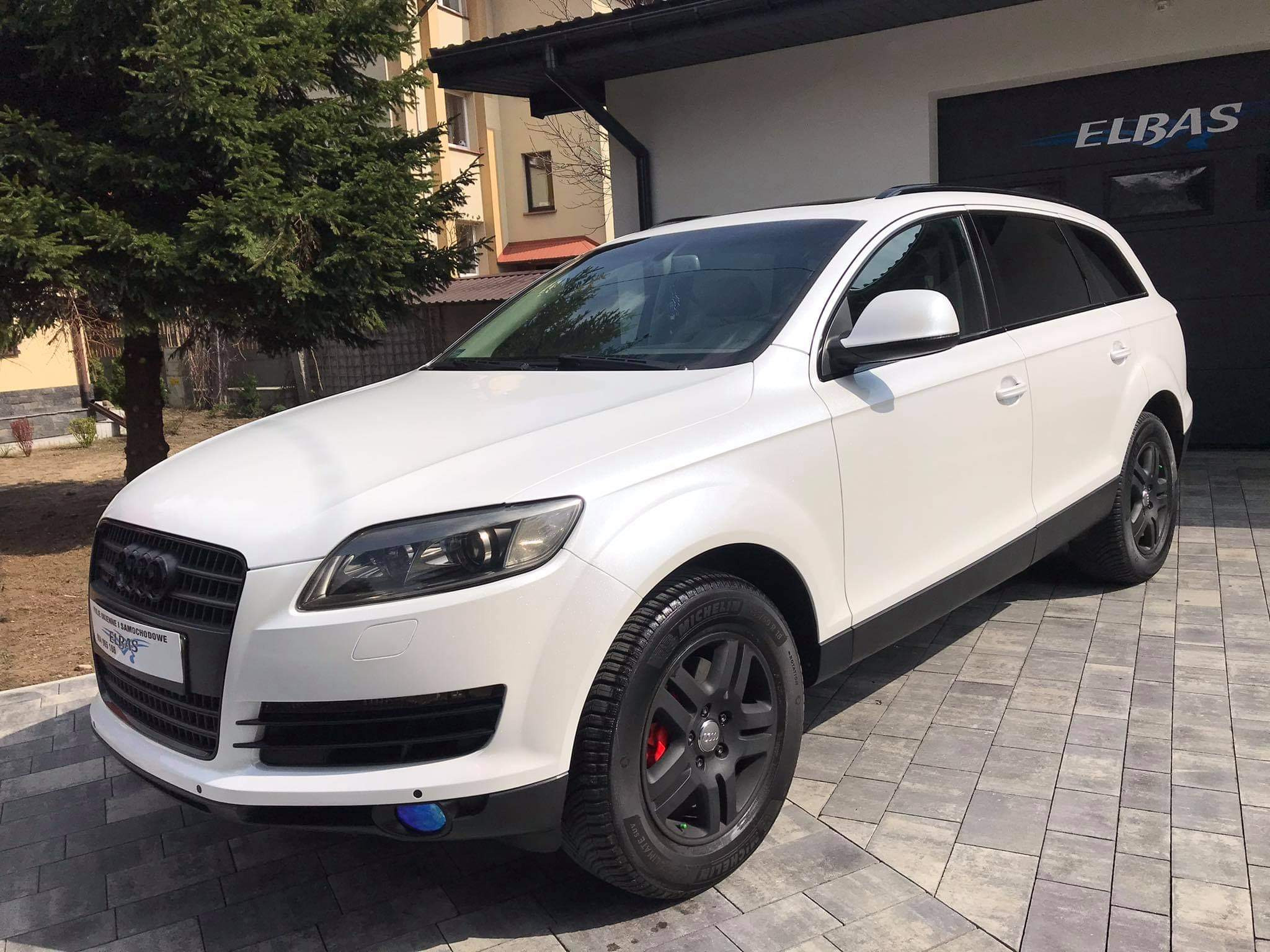Białe Audi Q7 z czarnymi felgami i zaczerwienionymi zaciskami hamulcowymi, zaparkowane na szarej kostce brukowej przed garażem z napisem 'ELBAS', widoczne przyciemnione tylne szyby.