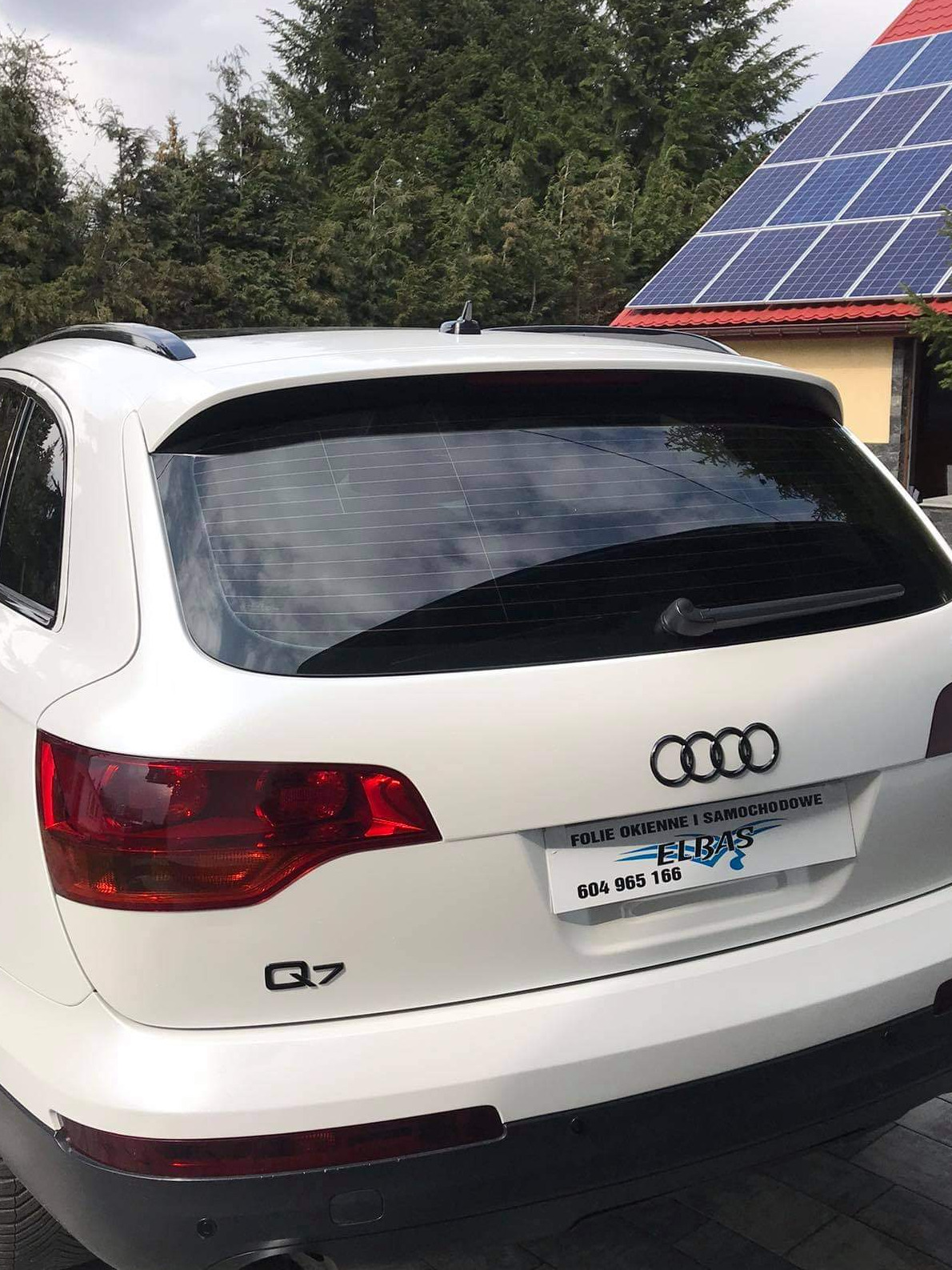 Białe Audi Q7 z przyciemnionymi tylnymi szybami, widoczne z tyłu na tle domu z panelami słonecznymi i drzewami.