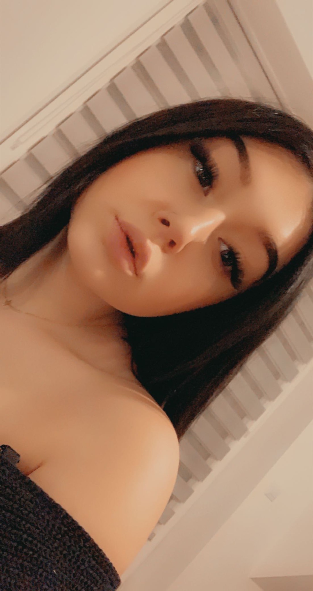 Portret młodej kobiety z długimi, ciemnymi włosami i delikatnym makijażem, skierowany w dół, z widocznym fragmentem czarnego swetra i złotym łańcuszkiem na szyi.