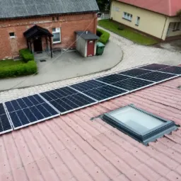 Rząd paneli fotowoltaicznych na czerwonej dachówce budynku, widoczne okno dachowe oraz budynki w tle.