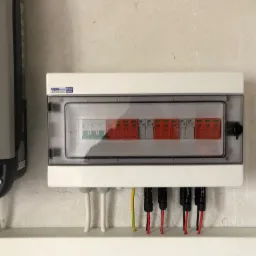 Szara skrzynka z bezpiecznikami i przezroczystym wieczkiem, zamontowana na teksturowanej ścianie obok szarego falownika z czarnymi elementami. Widoczne przewody wchodzące do skrzynki, w tym...