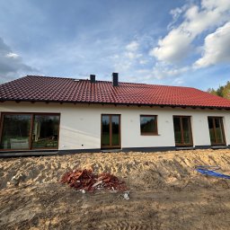 Kierownik Budowy/Inspektor Nadzoru Krystian Lindstaedt - Nowo wybudowany parterowy dom z czerwoną dachówką i brązowymi oknami, otoczony piaskiem i resztkami dachówek, z widocznymi rurami odprowadzającymi wodę.