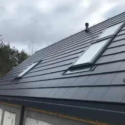 Czarny dach z dachówki ceramicznej z dwoma oknami dachowymi i kominkiem wentylacyjnym, widok z dołu na budynek w stanie surowym otwartym.