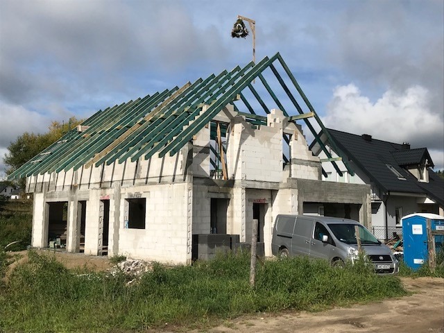 Budowa domu jednorodzinnego w stanie surowym otwartym: ściany z bloczków, zielona więźba dachowa, widoczny van i przenośna toaleta na działce.