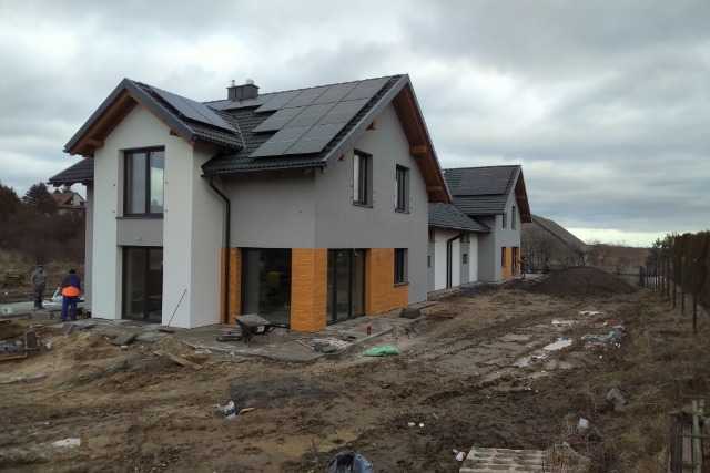 Dwa domy jednorodzinne w trakcie budowy, z panelami słonecznymi na dachu, widoczne prace ziemne i materiały budowlane na posesji w pochmurny dzień.