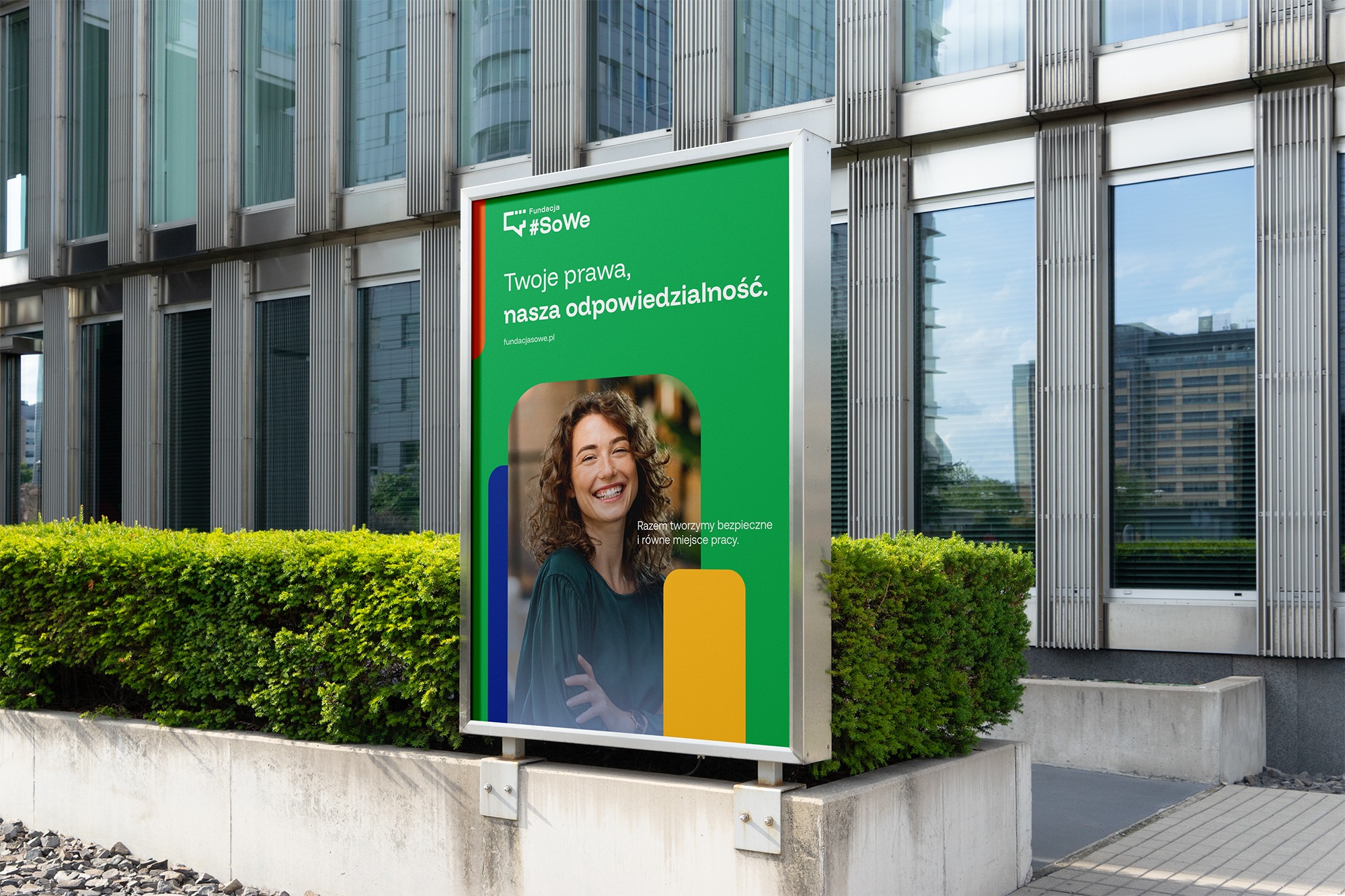 Billboard z zielonym tłem, hasłem #SoWe i uśmiechniętą kobietą na tle nowoczesnego budynku. Reklama fundacji na tle zieleni i szarej kostki brukowej.
