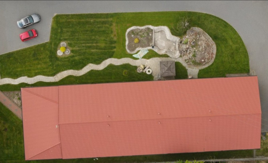 Widok z góry na budynek magazynowy z czerwonym dachem, otoczony zielenią, ścieżką i dwoma samochodami na parkingu.