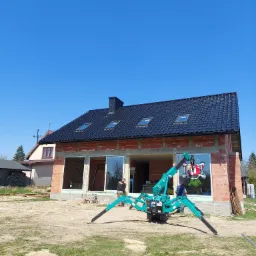 Turkusowy mini-dźwig pająk z przyssawkami podnosi dużą szybę okienną na placu budowy domu z czerwoną cegłą i czarnym dachem w słoneczny dzień.