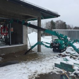 Warsaw Bridge Sp. z o.o. - Mały, zielony dźwig pająk z przyssawkami montuje okno w surowym stanie budynku, na zewnątrz leży śnieg.