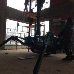 Warsaw Bridge Sp. z o.o. - Montaż okien za pomocą małego dźwigu gąsienicowego w surowym stanie budynku, pracownicy na rusztowaniu w tle, widoczne przyssawki do szkła.